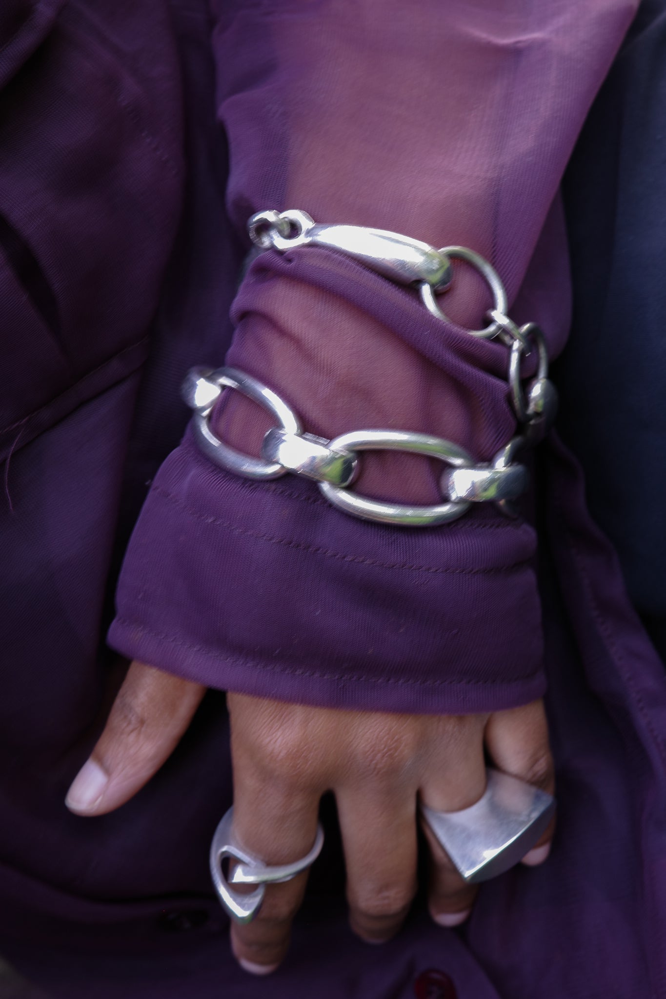 Silver chain bracelet and rings on a person wearing a purple garment.